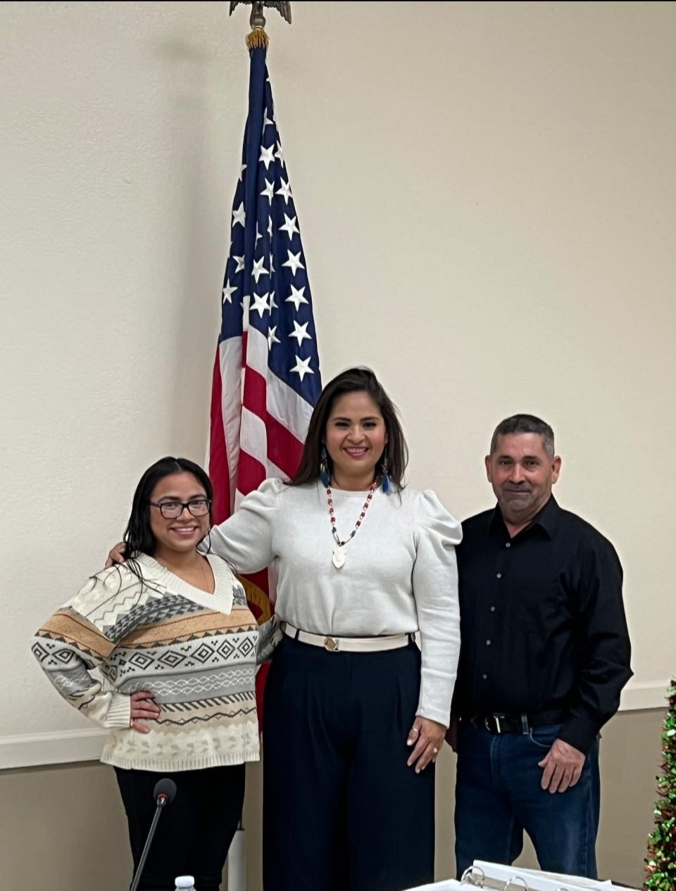 Three people stand indoors in front of a U.S. flag, smiling.
