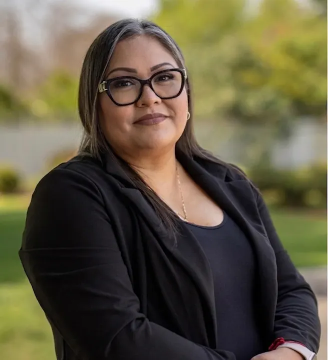 A woman with glasses and long hair is standing outdoors, wearing a black blazer, with a soft smile against a green background.