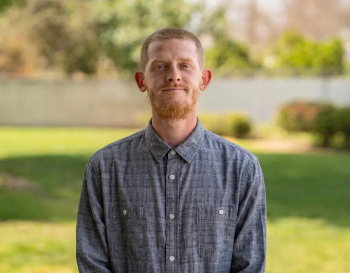 A person with a beard stands in a green outdoor setting, wearing a button-up shirt and smiling.