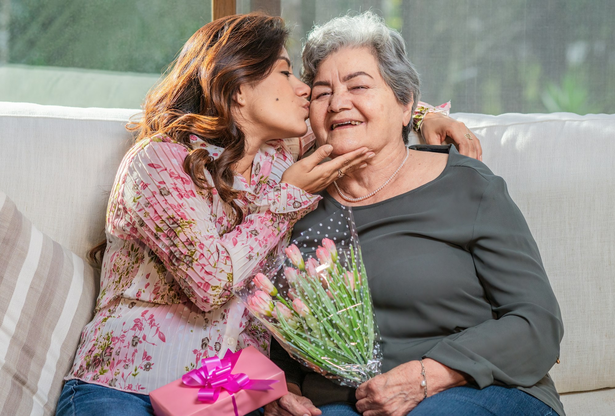 A young woman kisses an older woman, holding flowers and a gift, seated on a sofa.