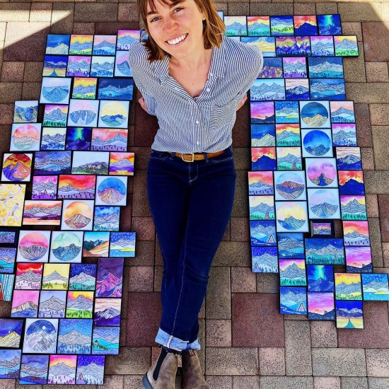 A happy person surrounded by colorful paintings, likely their own artwork, laid out on the ground.