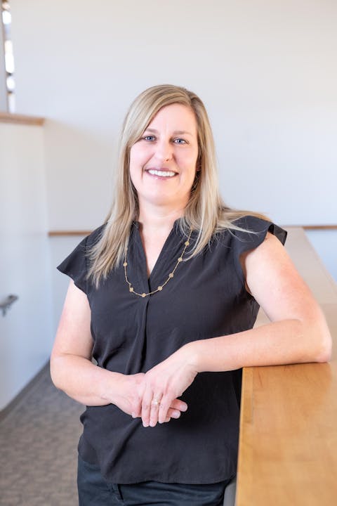 Amy KIng PA-C smiling, with long blonde hair, wearing a black top and gold necklace, stands with her hand on a wooden surface.