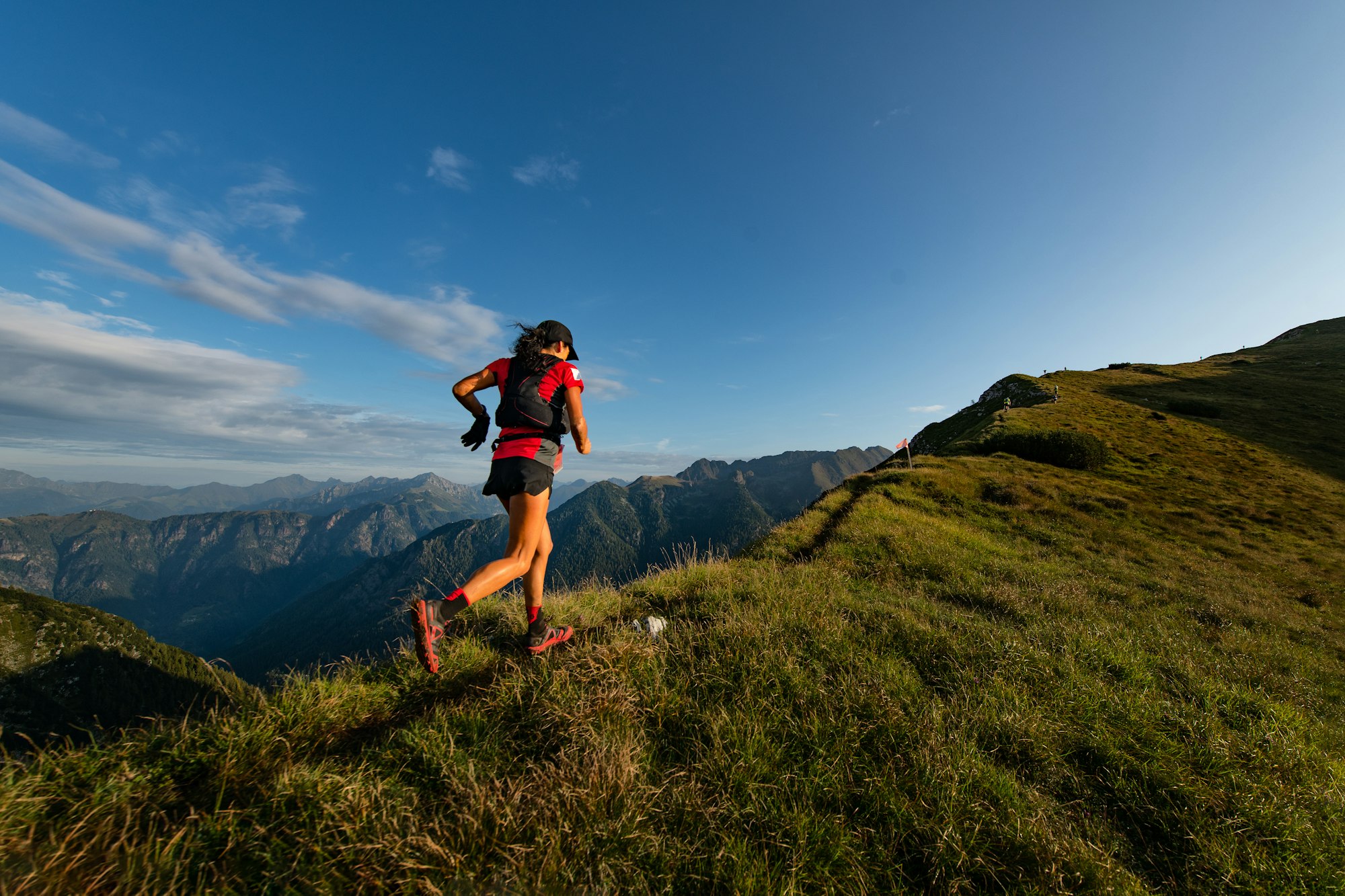 A person trail running along a grassy hillside with a mountain landscape in the background under a clear blue sky.