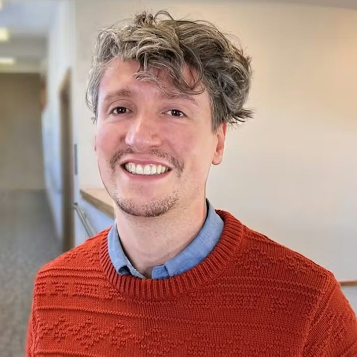 A smiling person in an orange sweater with a patterned design stands in a well-lit indoor space.