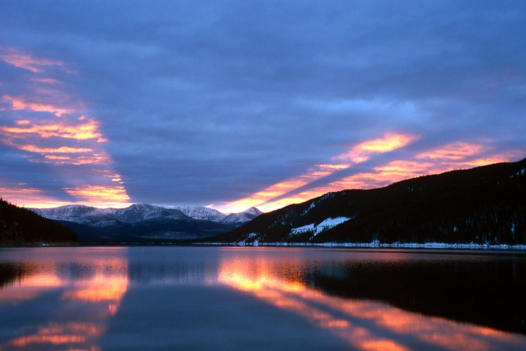 A serene lake at dusk with reflecting sunset skies and snow-capped mountains in the background.