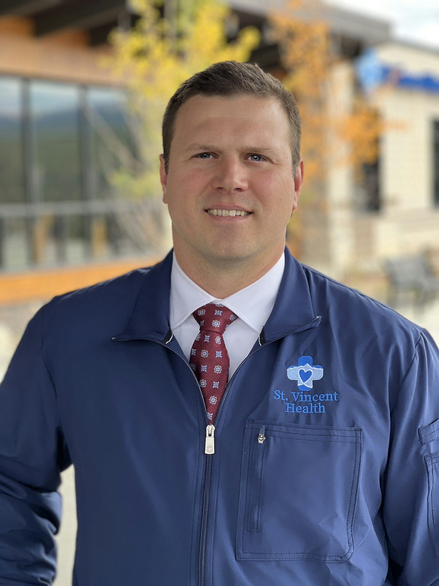 Bubba Bartlett in a navy jacket with "St. Vincent Health" logo, wearing a red tie, outdoors near a building with blurred background.
