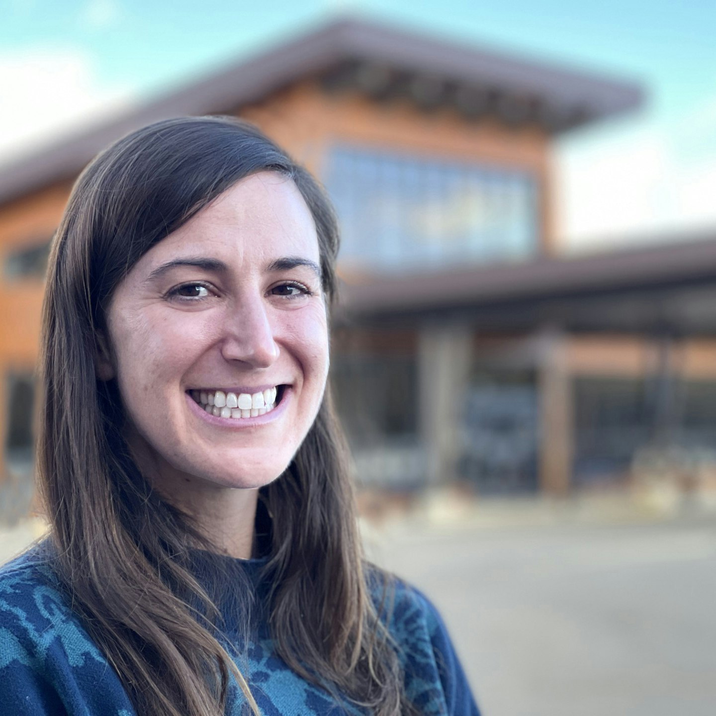 A smiling woman stands in front of a modern building with large windows, showcasing a casual yet friendly demeanor.