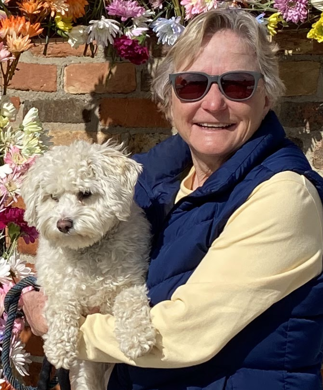 Sarah Savage wearing sunglasses holding her fluffy white dog in front of a brick wall with colorful flowers.