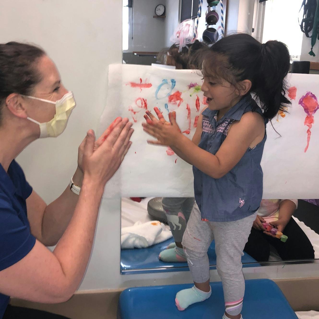 A child paints on a vertical surface while an adult kneels nearby, both engaged in the activity.