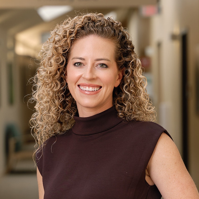 Alexandra Kovar, MD smiling, with curly hair, wearing a sleeveless dark top, stands in a softly lit hallway.