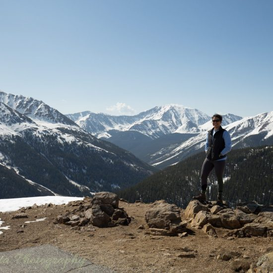 Person standing on a mountainous overlook with snowy peaks in the background.