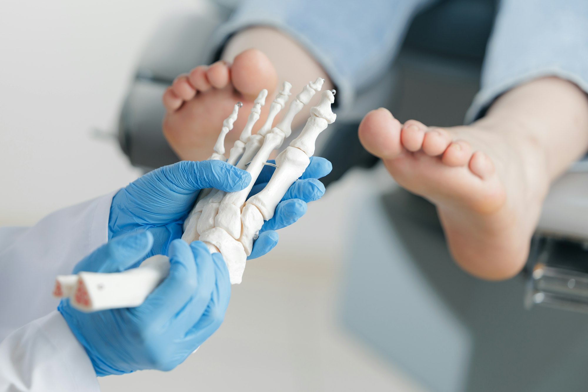 A medical professional holds a foot skeleton model while examining a patient's feet in a clinic setting.