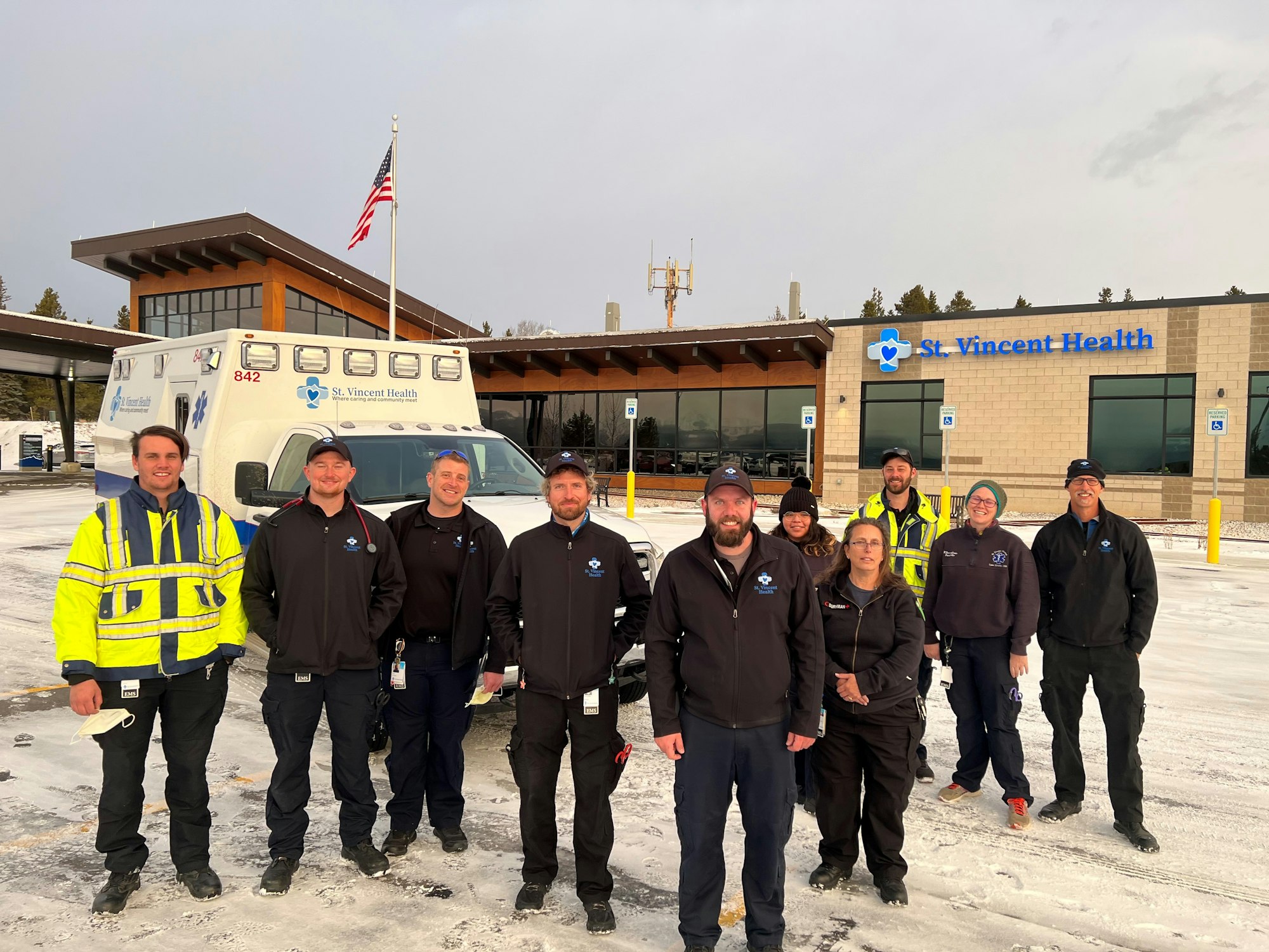 A group of St. Vincent Health first responders standing in front of an ambulance and St. Vincent Health building in a snowy area.