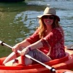 A smiling person kayaking, wearing a hat and sunglasses, on a sunny day.