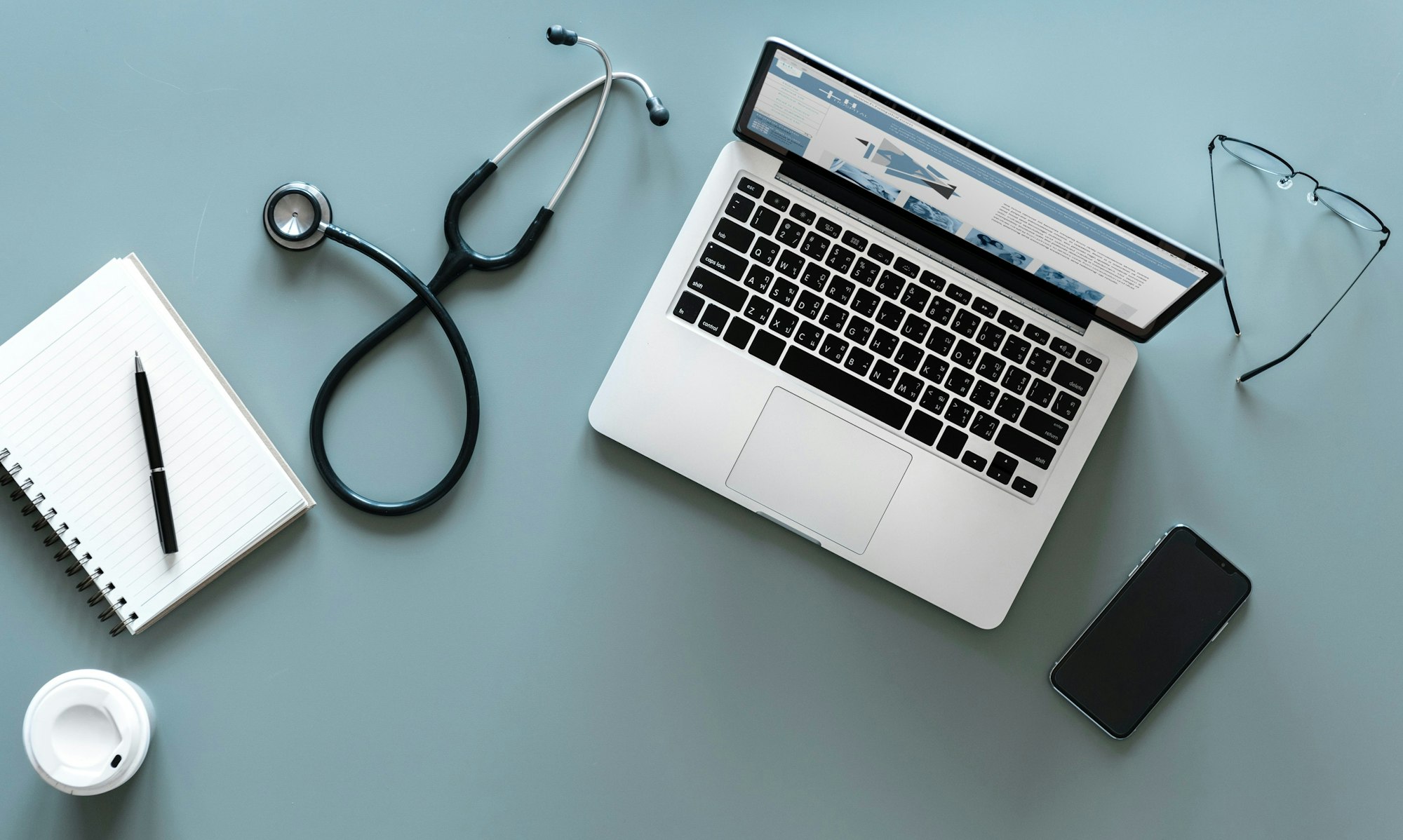 A laptop, stethoscope, notebook, pen, glasses, smartphone, and coffee cup on a blue surface.
