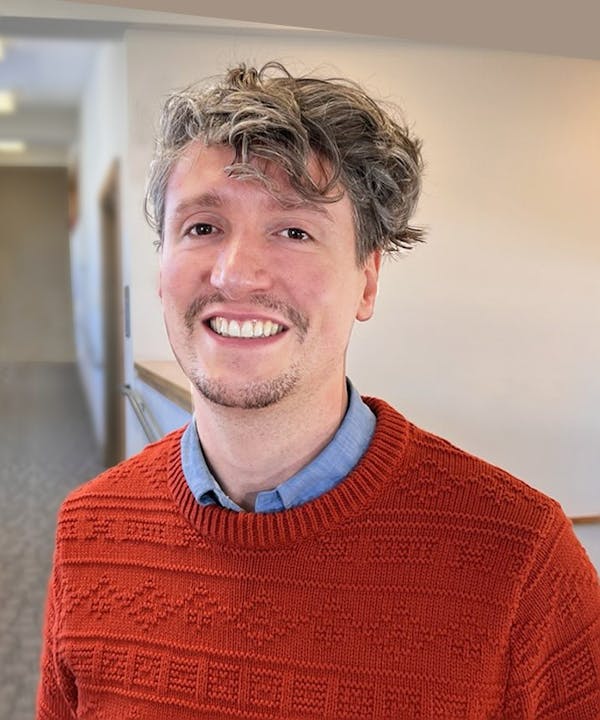 Dr. Tsambikos smiling, wearing an orange sweater over a blue shirt, standing in a hallway.