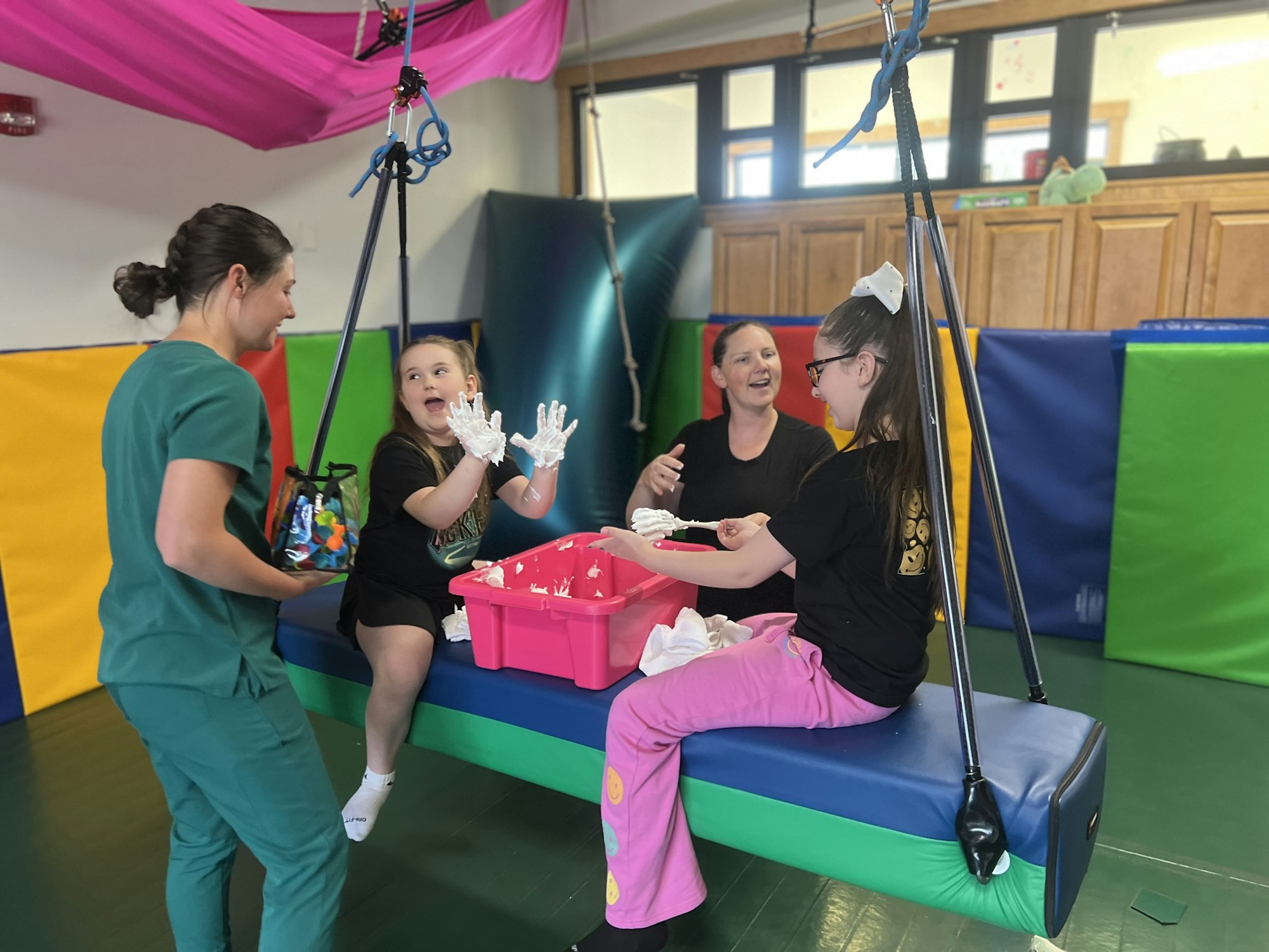Two children playing on a suspended swing with colorful mats around, assisted by two adults.