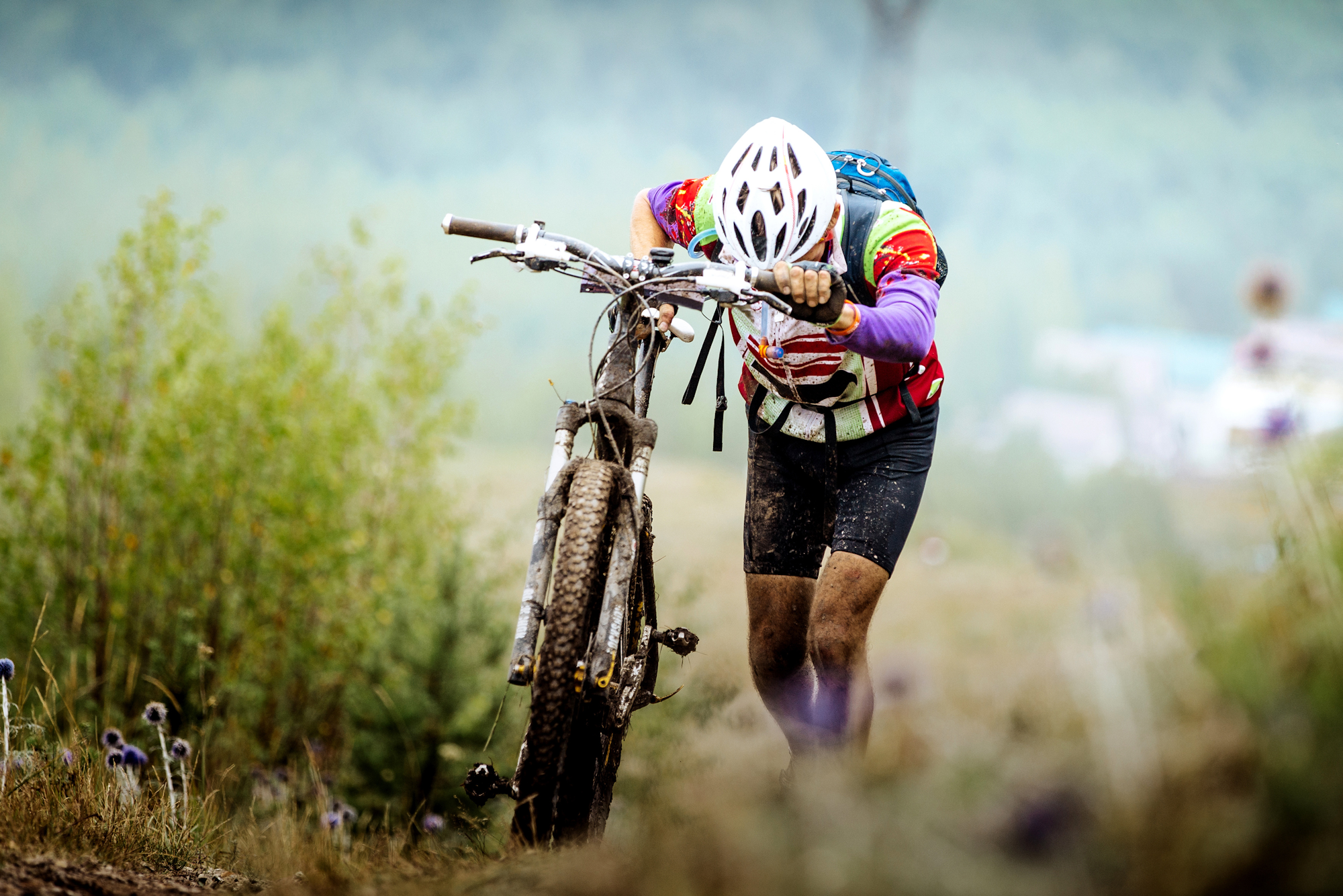 A cyclist in a helmet pushes a muddy mountain bike uphill through a grassy, misty landscape.
