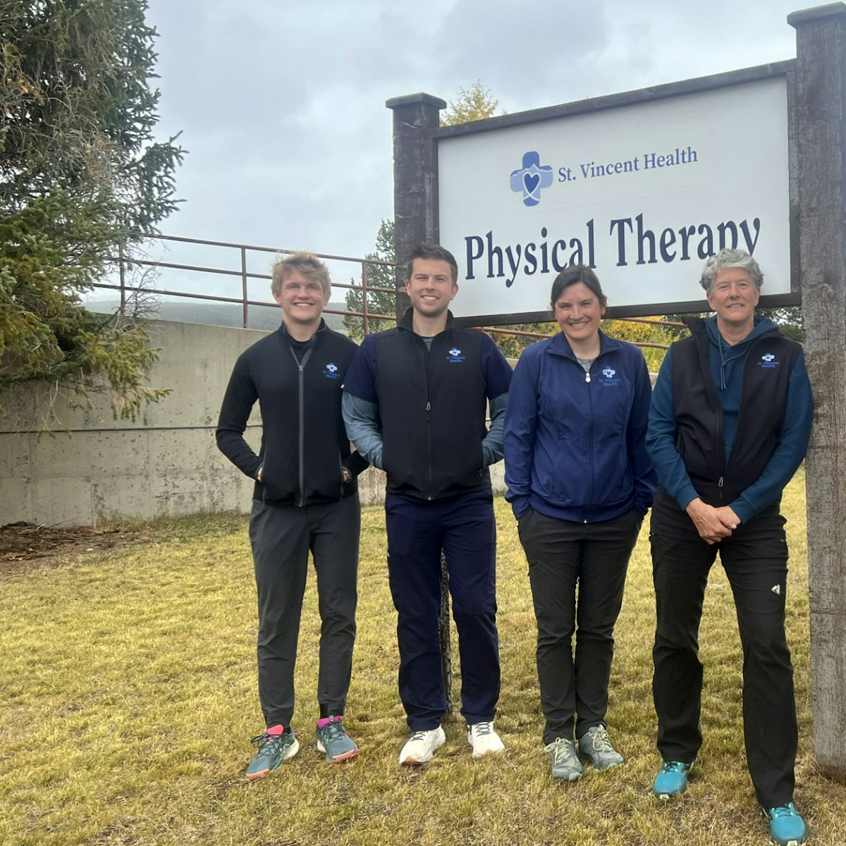 Four physical therapists standing in logoed clothing outside near a "St. Vincent Health Physical Therapy" sign on a grassy area.