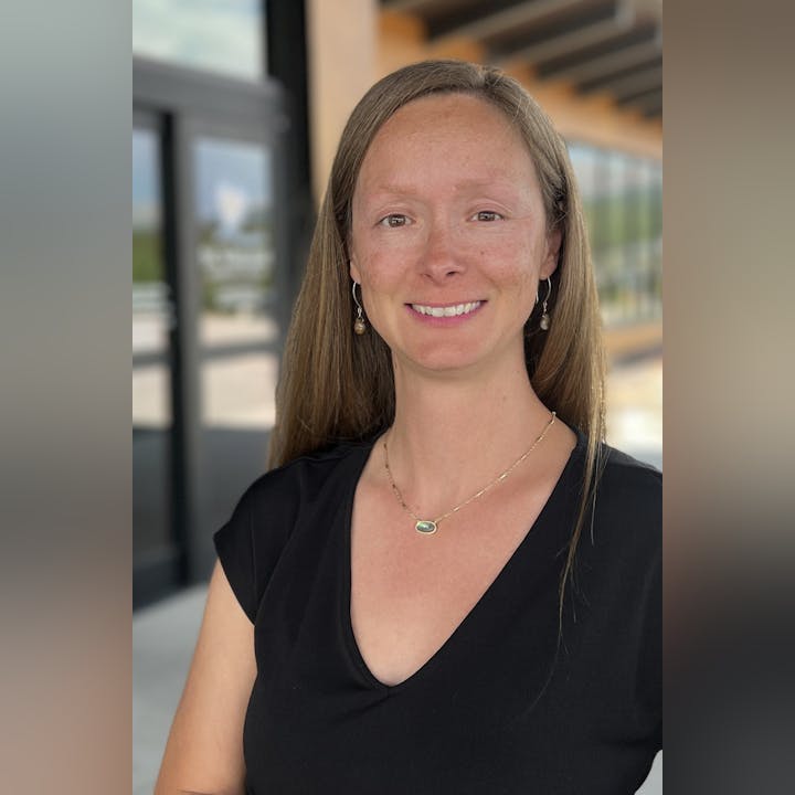 Joanie Yapkowitz wearing a black top, standing outdoors with a blurred background of windows and a door.