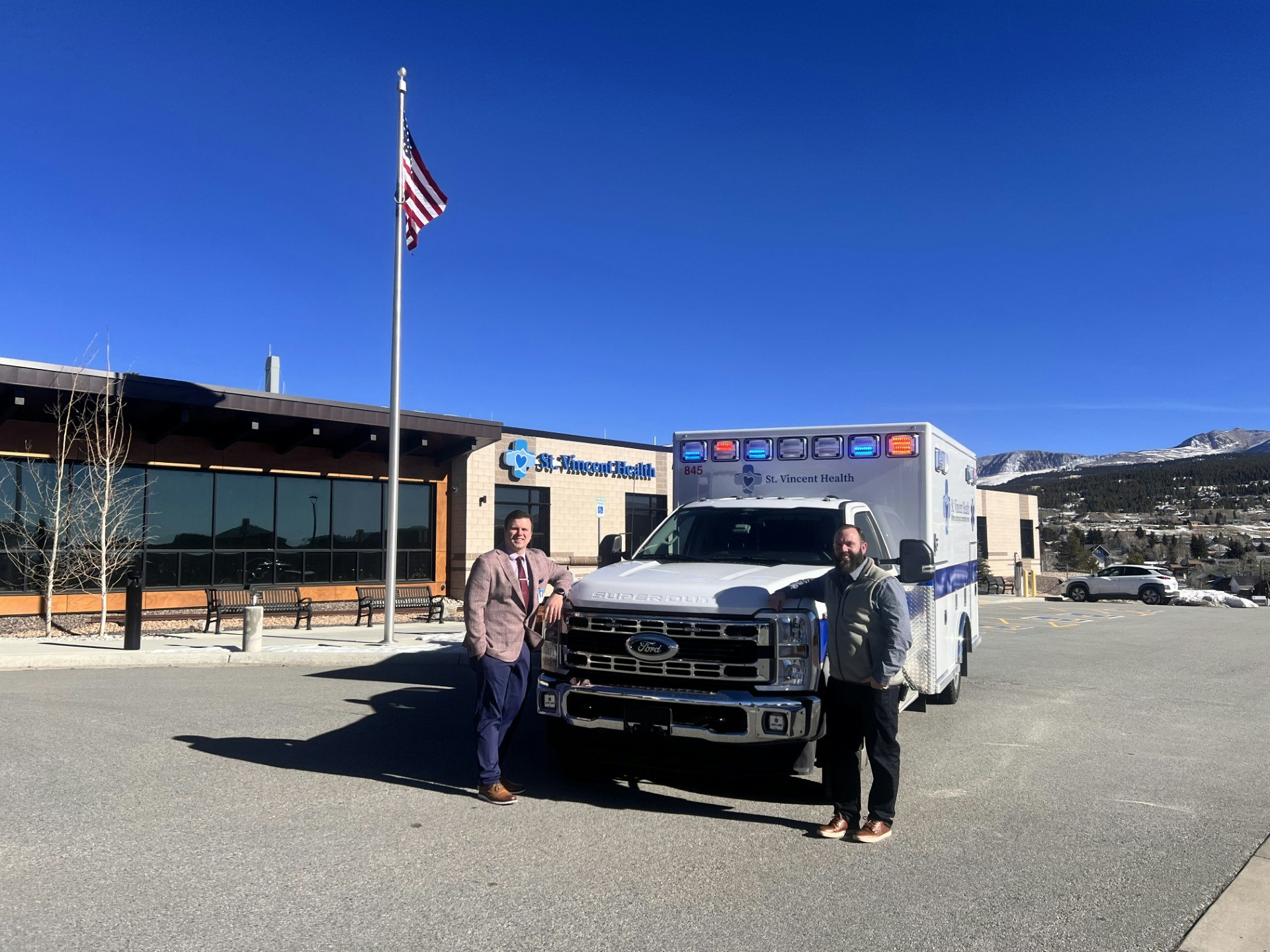 CEO, Bubba Bartlett and Ambulance Director, Jeremiah Grantham stand with the new 2025 ambulance outside a St. Vincent Health facility, with an American flag and clear blue sky in the background.