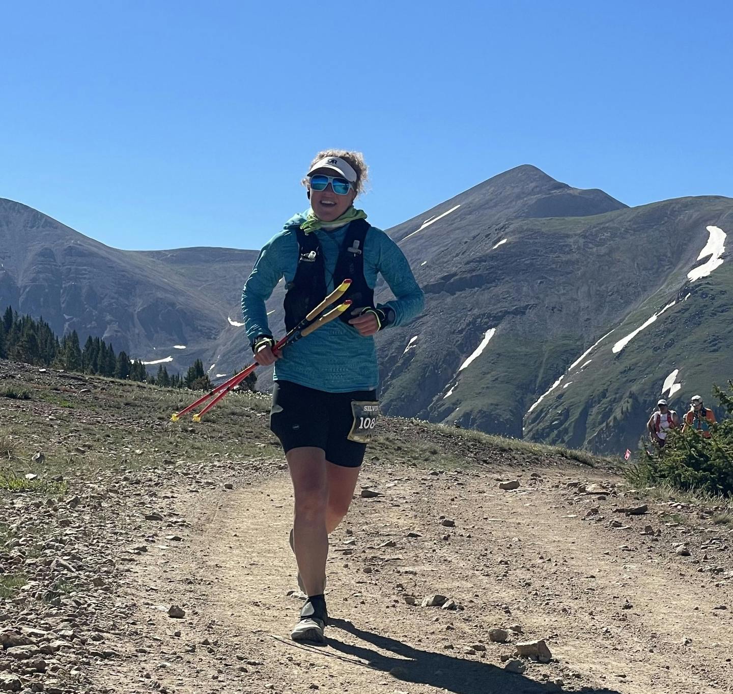 Kelli McCall in a blue jacket is navigating a mountainous trail under a clear sky, using trekking poles.
