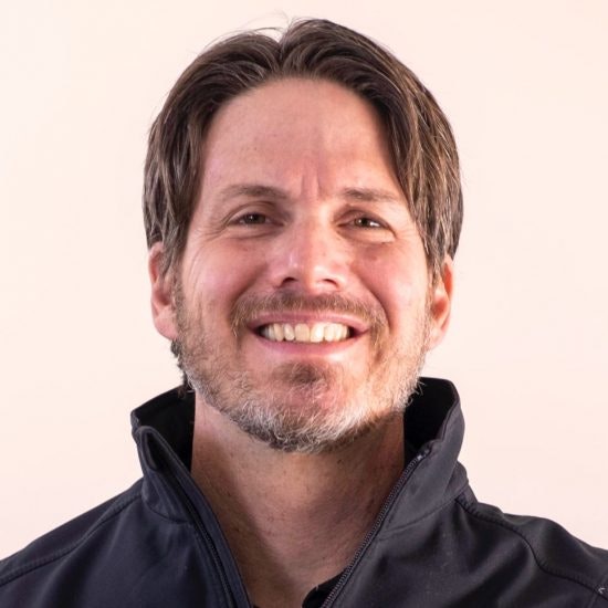 Smiling man with stubble wearing a black jacket against a pale background.