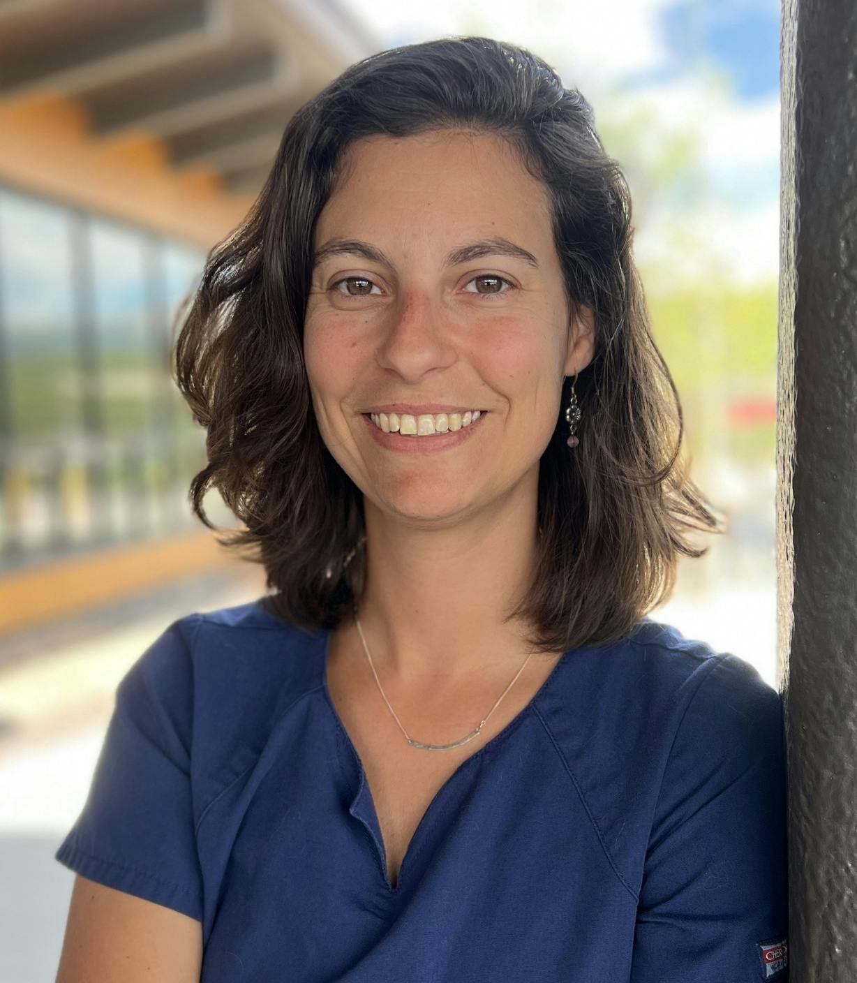 Lindsy Coon, smiling in blue scrubs leans against a pillar outside, with a blurred building and sky in the background.
