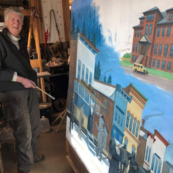 Person with a painting depicting a street and buildings, smiling, in a workshop with tools.