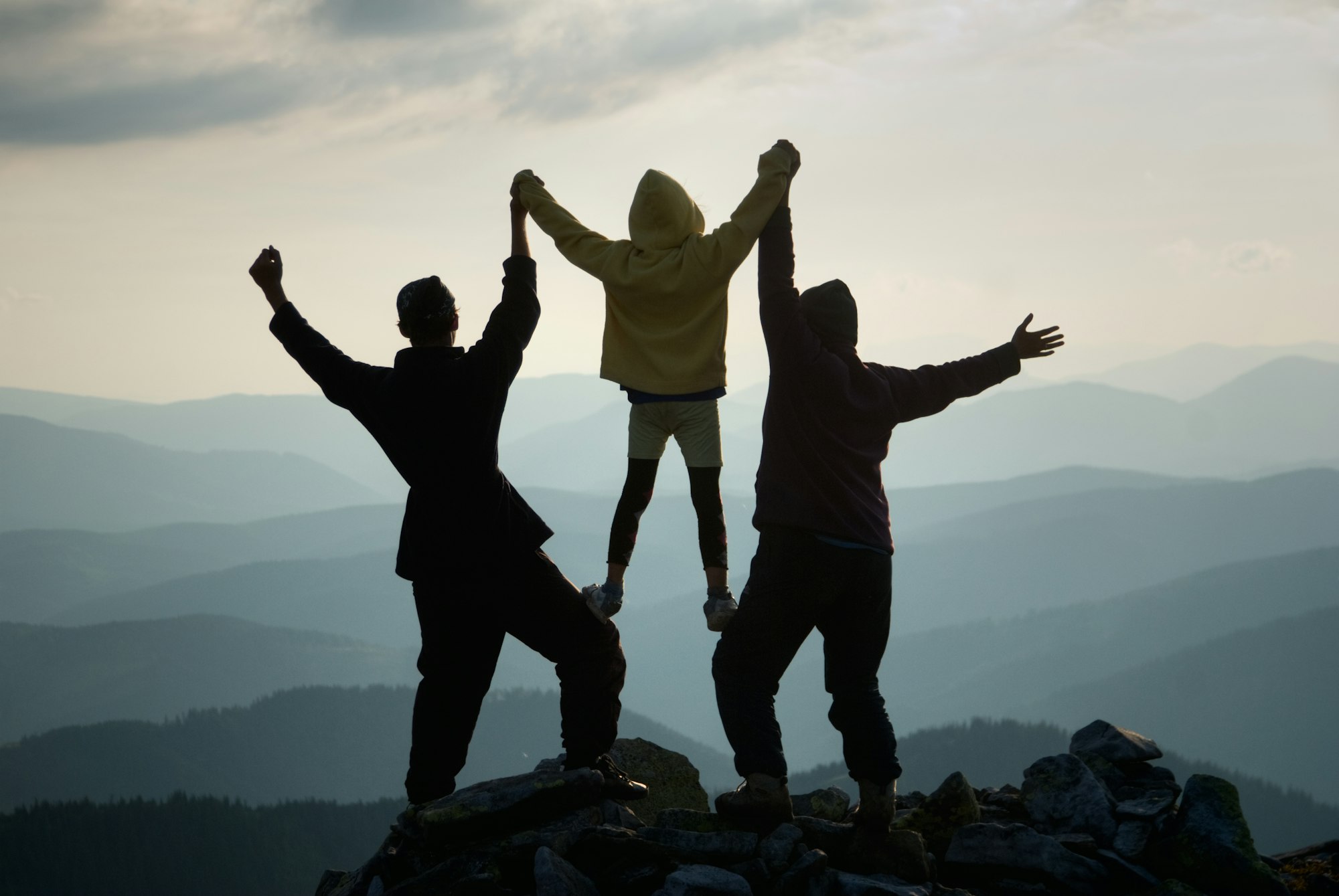 Three people stand on a mountain peak, arms raised, celebrating with a scenic view behind them.