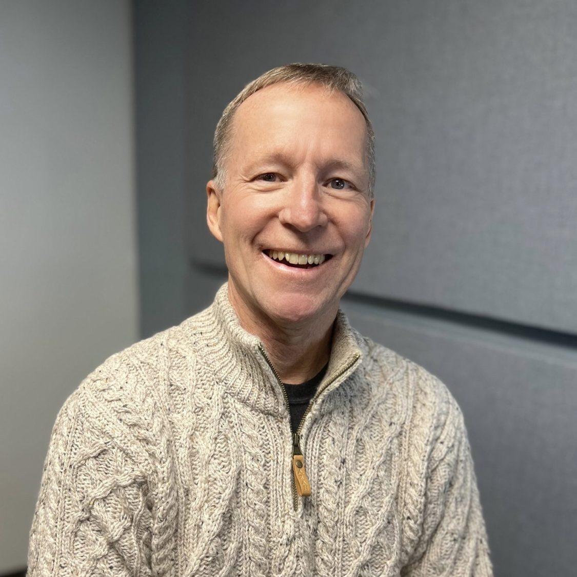 Jay Coon smiling in a cable-knit sweater against a grey background.