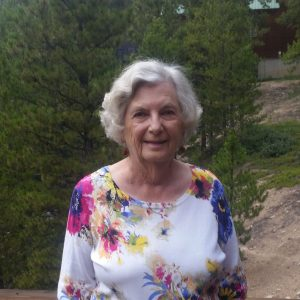 A smiling elderly lady in a floral shirt standing in front of trees.