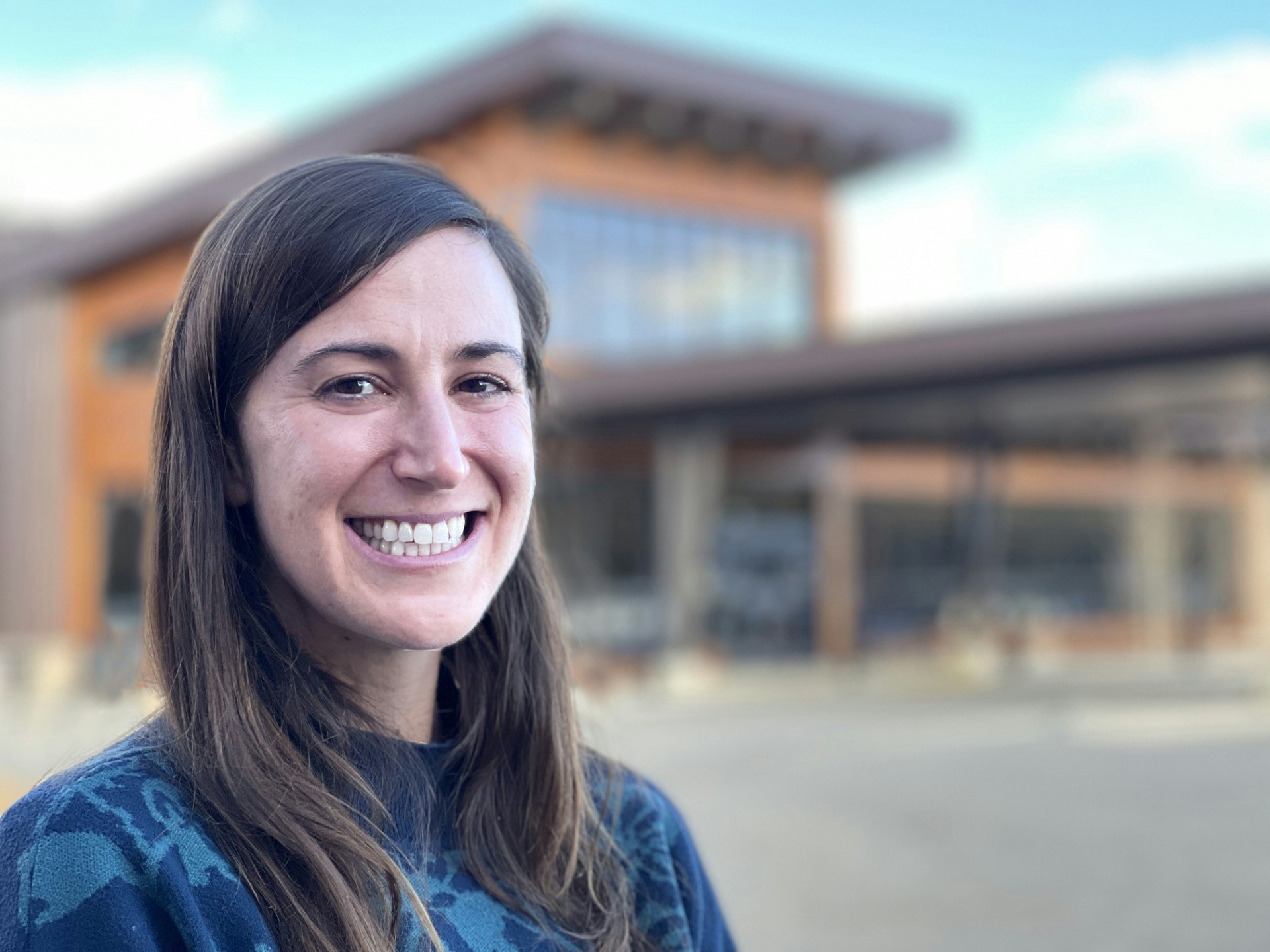 A smiling woman stands outdoors in front of a modern building, showcasing a cheerful expression and casual attire.