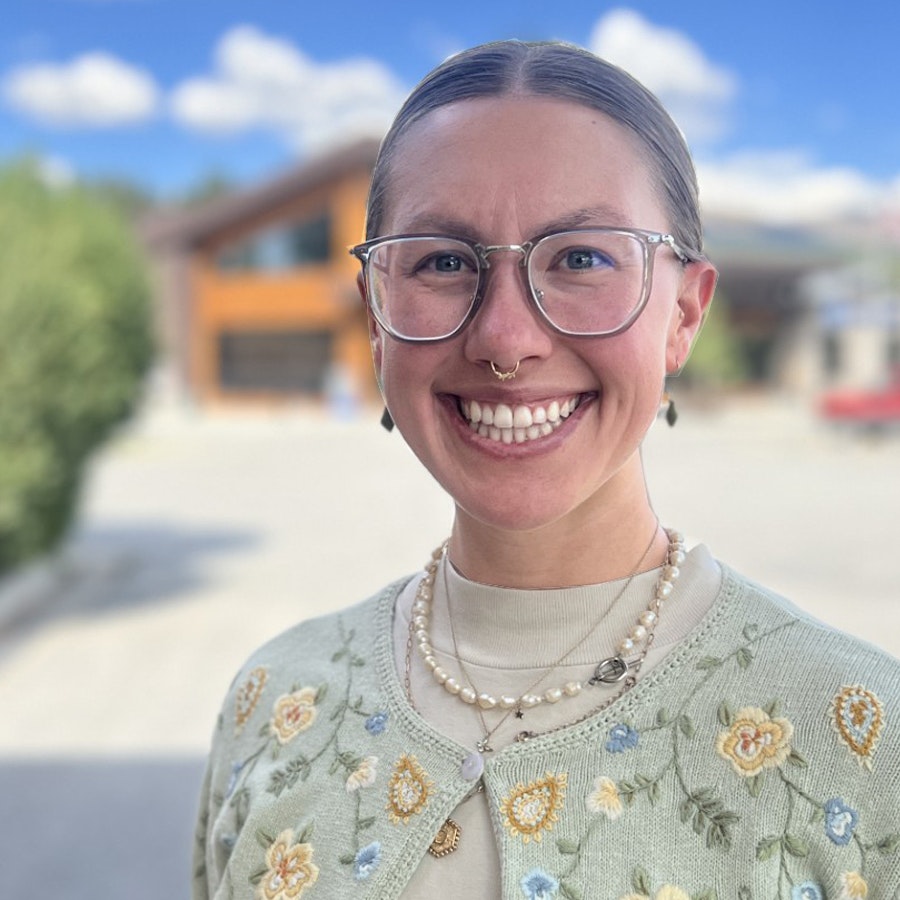 A smiling person with glasses and a septum piercing wears a floral cardigan, standing outdoors with a blurred background.