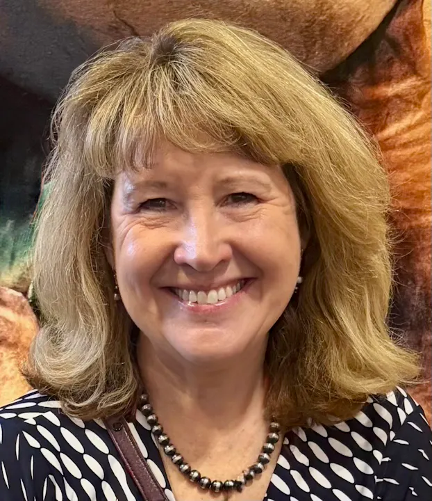 Karen Casey smiling with blonde hair, wearing a polka dot blouse and a beaded necklace, stands in front of a colorful background.
