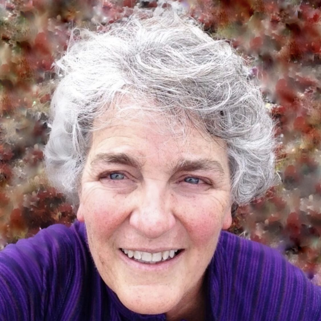 A smiling person with gray hair and a purple top against a blurred background.