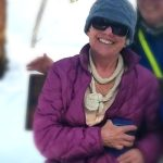 A smiling person wearing sunglasses, a purple jacket, and a cap, possibly outdoors.