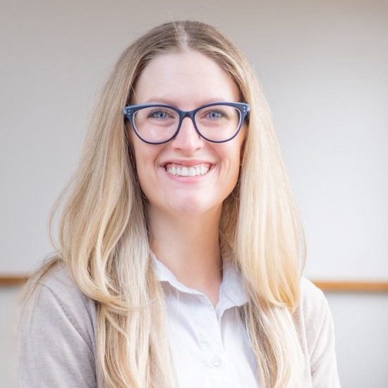 A smiling woman with blond hair, blue framed glasses, and a white shirt against a neutral background.
