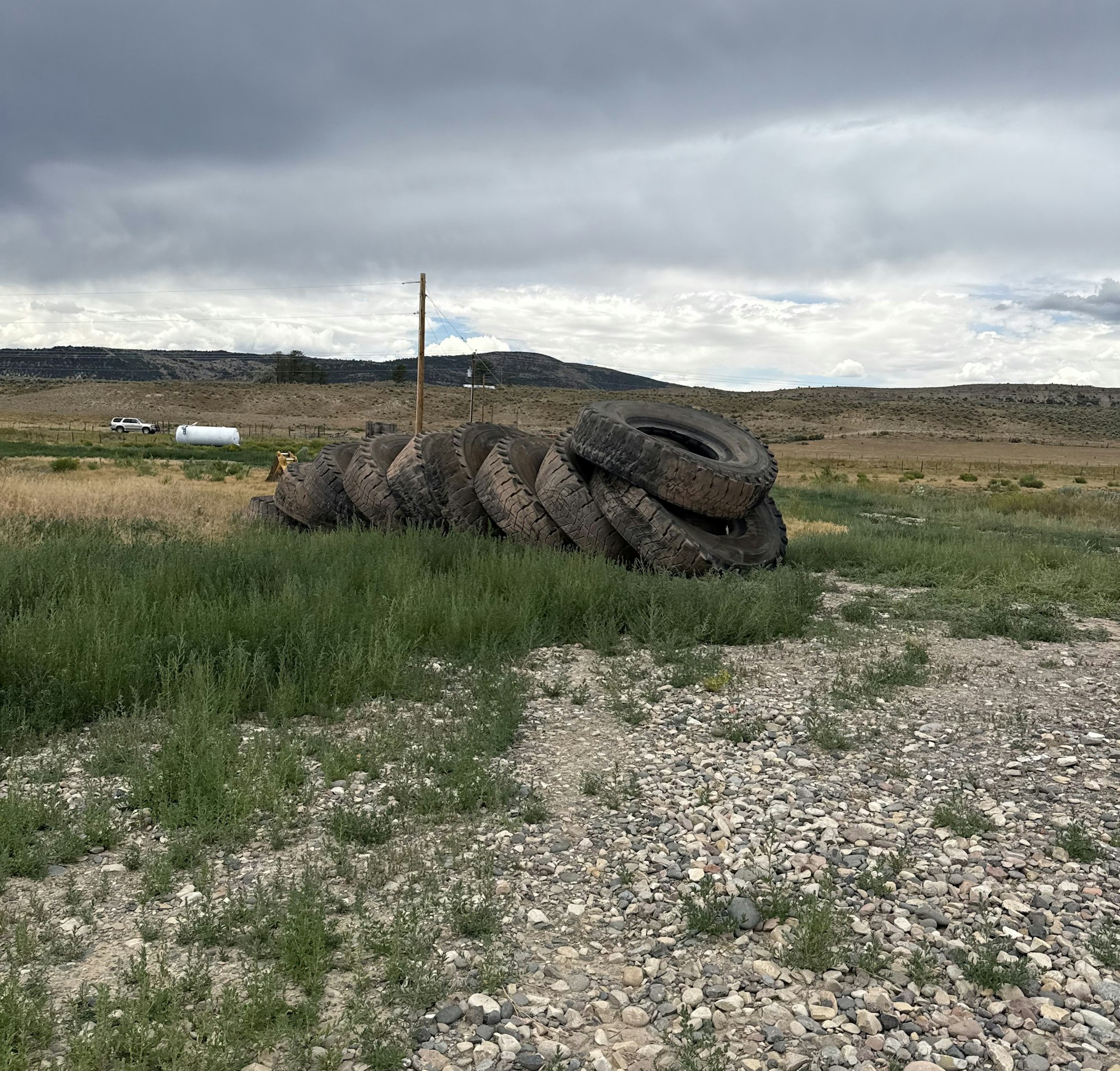 A stack of large tires lies on a grassy area with a rocky path, set against a backdrop of hills and cloudy skies.