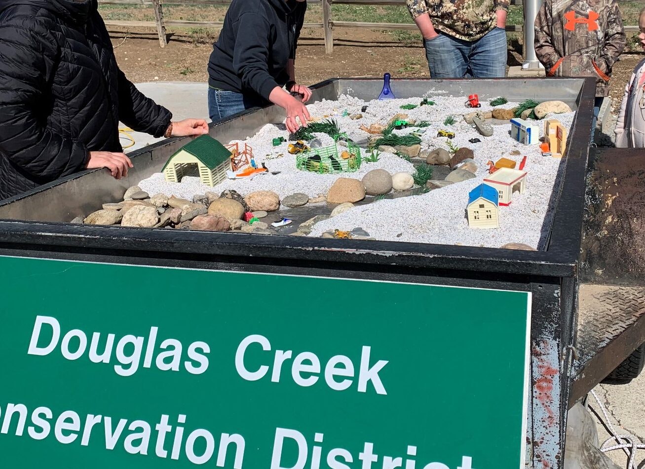 A tabletop model with rocks and miniature structures, surrounded by people, labeled "Douglas Creek Conservation District."