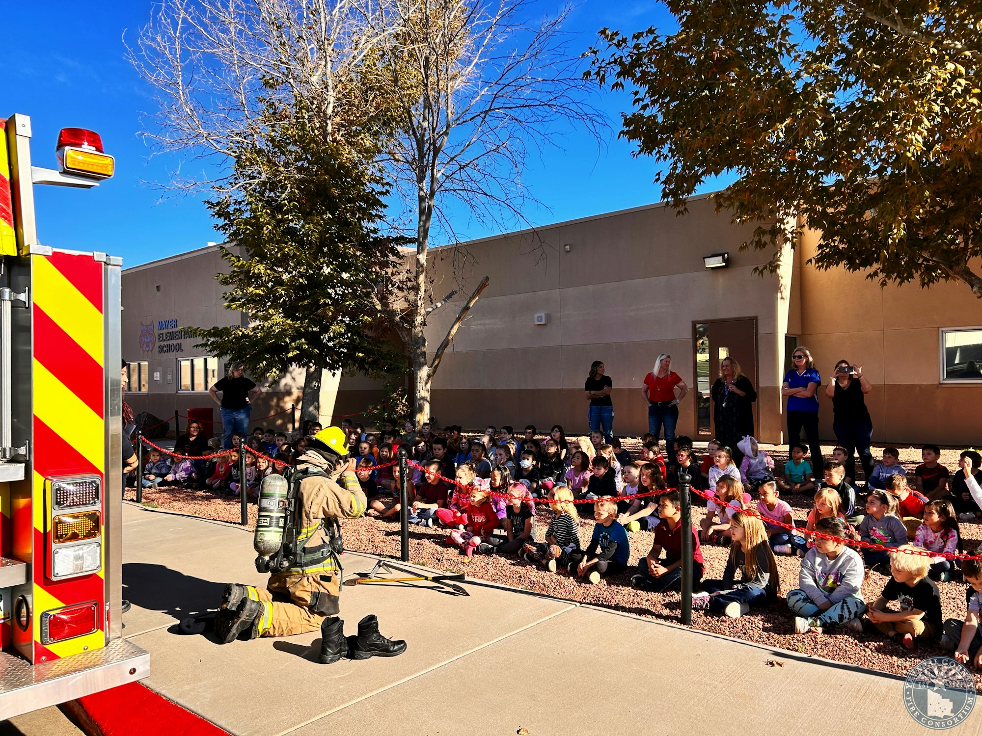 Firefighter kneels beside fire truck, demonstrating to children seated outside a school building on a sunny day.