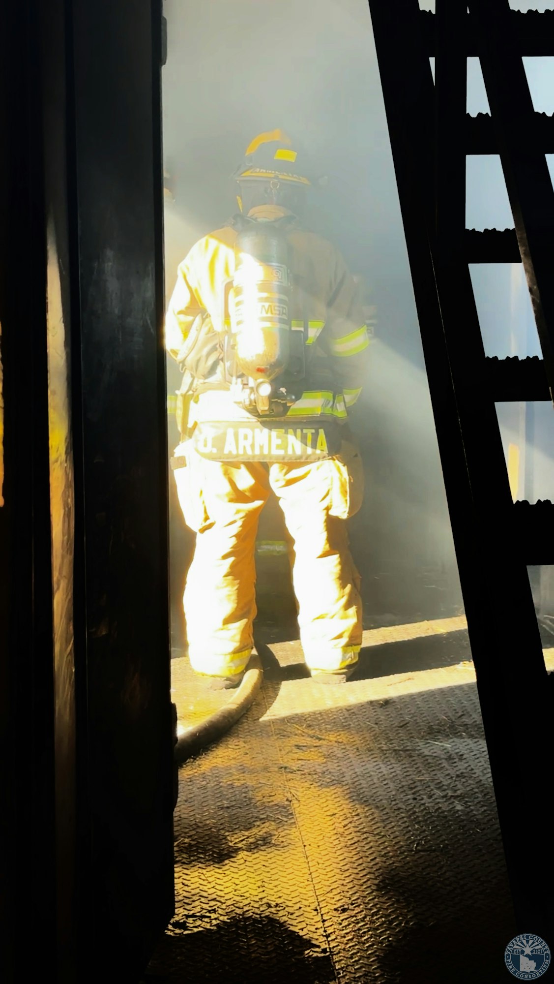 A firefighter in full gear stands in a smoky environment, illuminated by light, next to a ladder.
