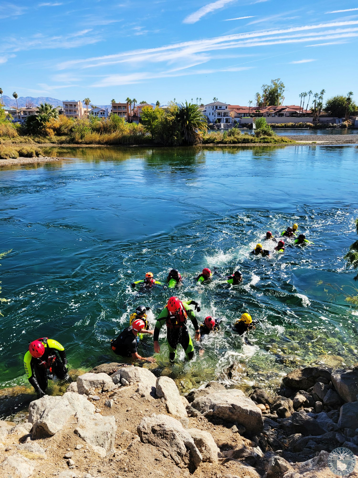 People in helmets and wetsuits emerge from a river onto a rocky shore, with buildings and trees in the background.