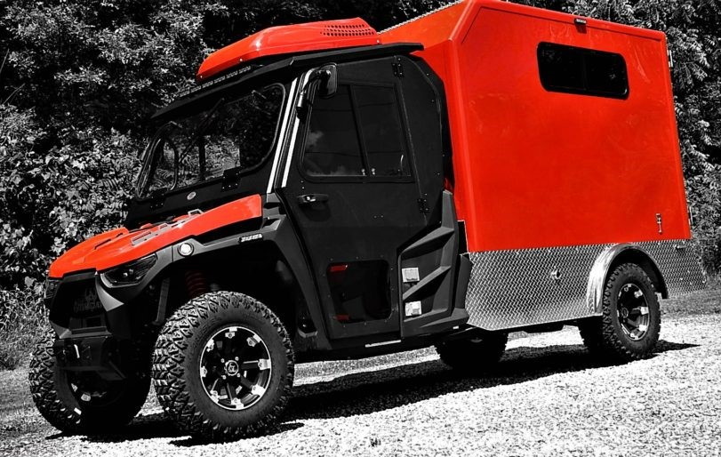A red and black off-road vehicle with a boxy rear cab, parked on gravel with trees in the background.