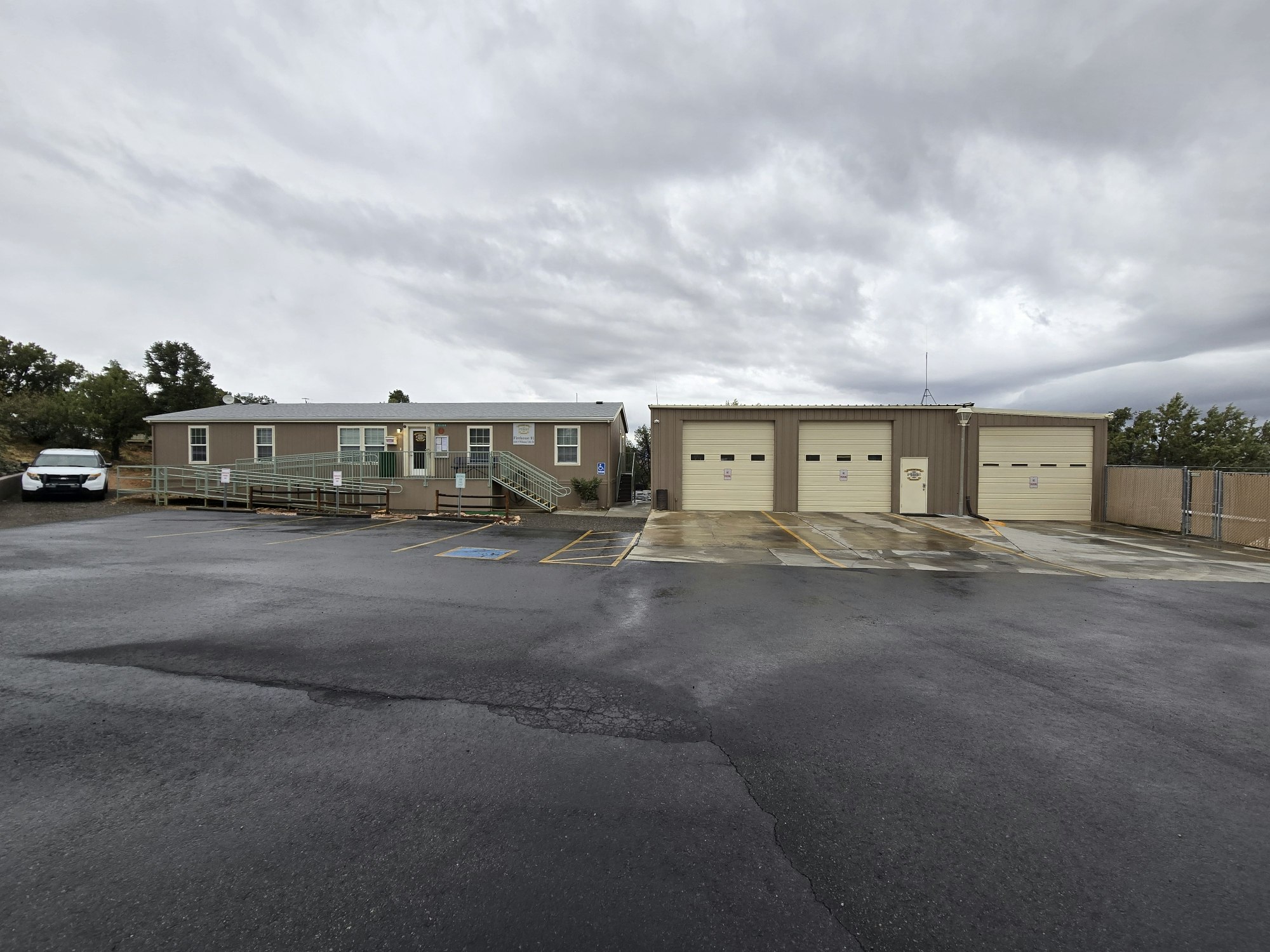 A small building with a ramp, an adjacent garage with three bays, and a parked car in a paved lot under a cloudy sky.