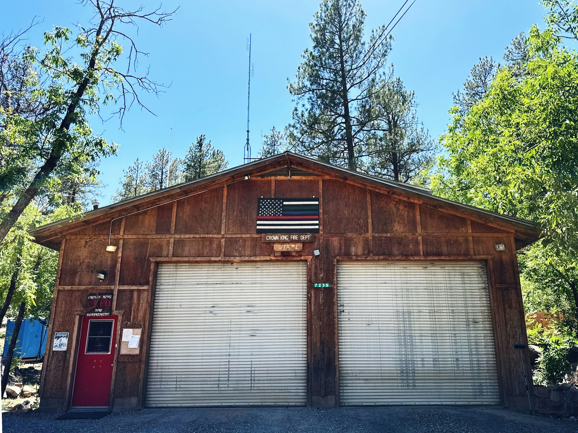 A wooden fire station building with a U.S. flag, trees around, two garage doors, and a red entry door.