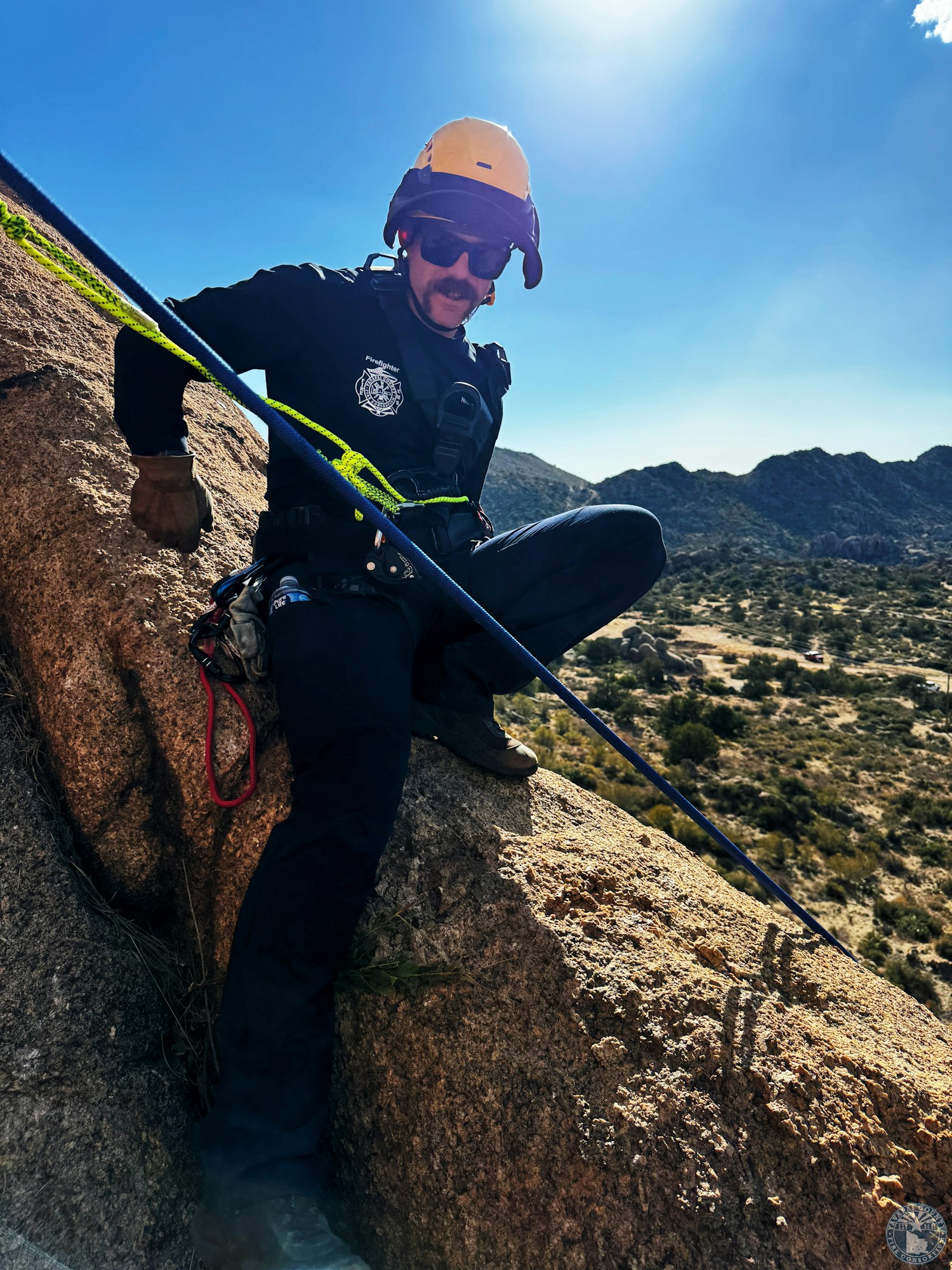 A person in climbing gear rappels down a rocky cliff in a sunny, mountainous landscape.