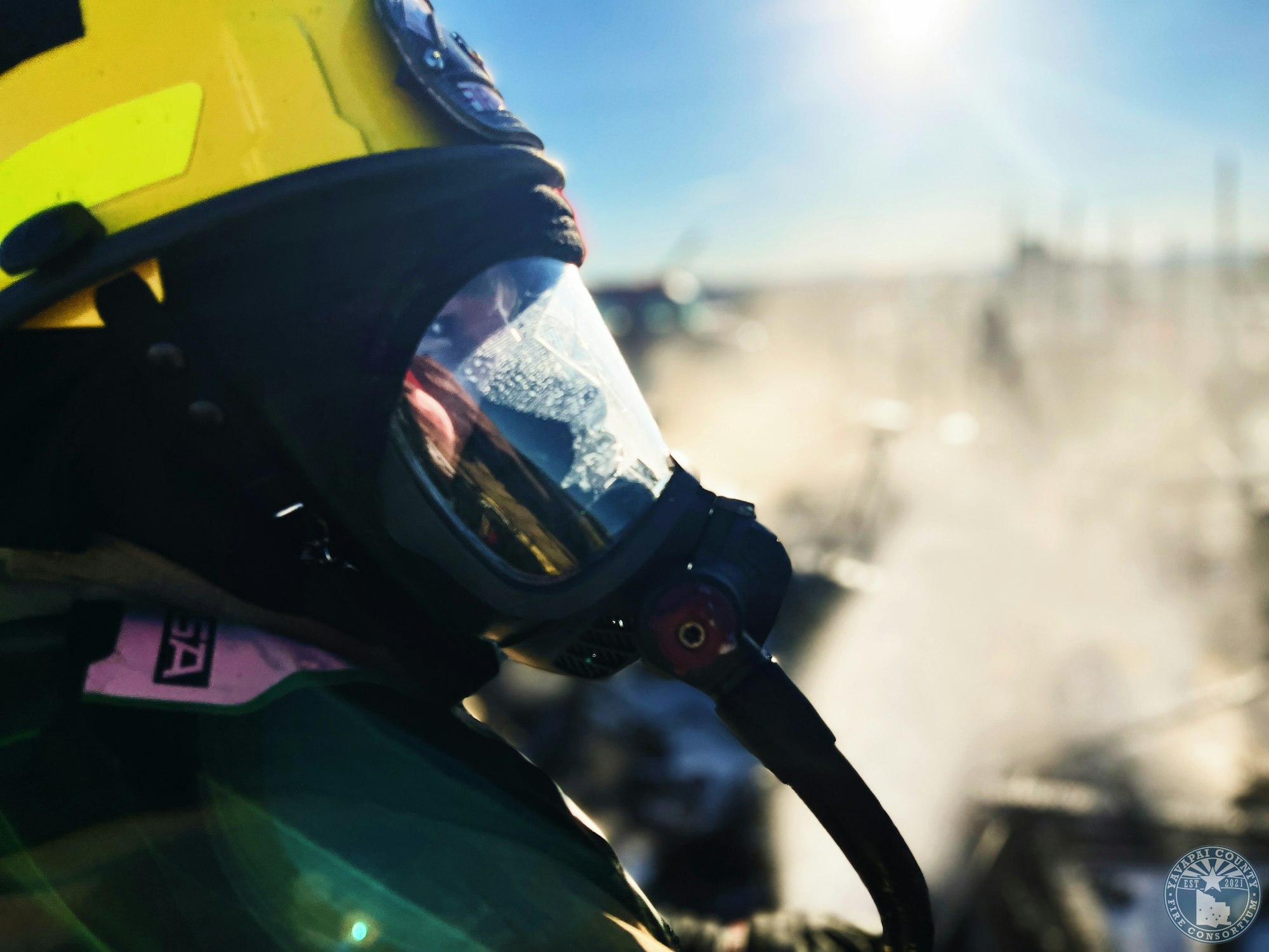 Firefighter in full gear and mask, facing a bright, smoky scene.