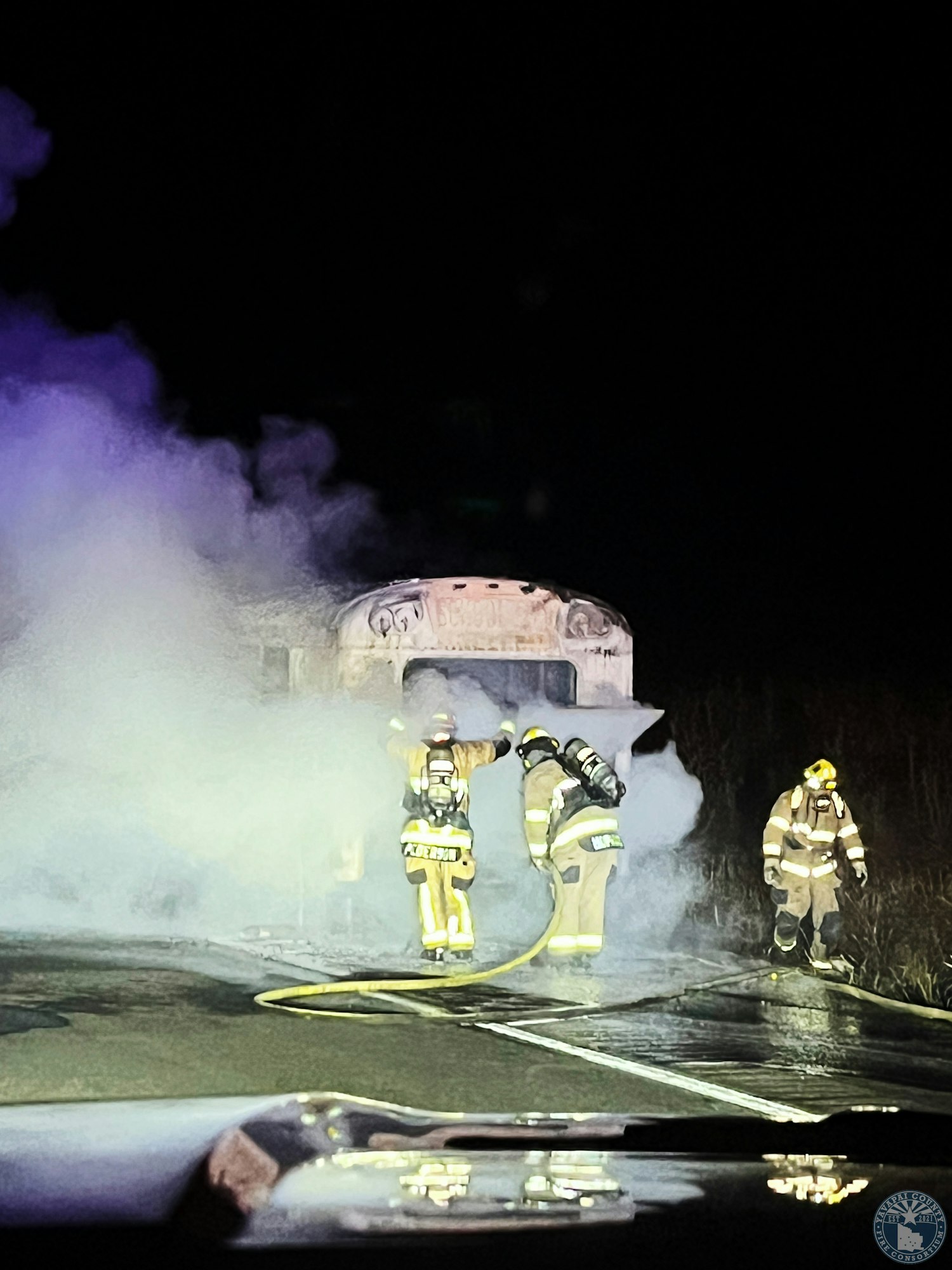Firefighters extinguishing a vehicle fire on a road at night, surrounded by smoke.
