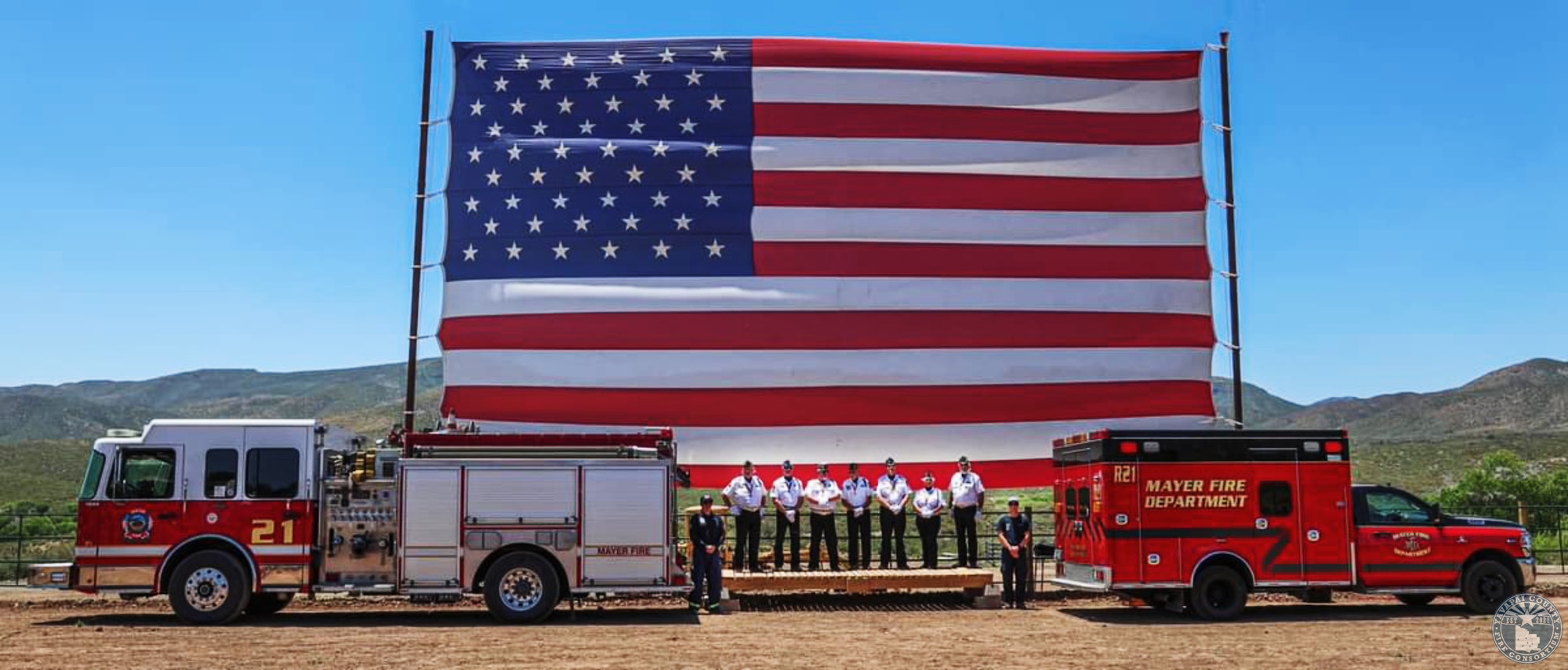 Firetruck, ambulance, and personnel in front of a large American flag in a scenic outdoor setting.